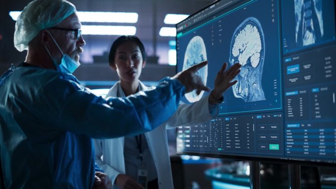 医院研究实验室会议:亚洲女性神经科学家与高加索男性神经外科医生讨论电视屏幕上的CT脑部扫描。穿着手术
