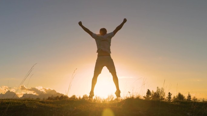 镜头光晕:日出时，一个年轻人在长满草的山顶上举起双臂跳跃
