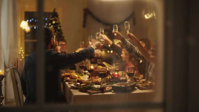 拉近镜头，透过窗户，老人和年轻的家庭成员举起并碰杯，在家里庆祝节日的圣诞之夜。亲戚们正在享用火鸡大餐