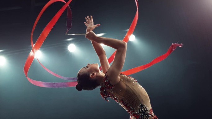 灯光大舞台上的现代艺术体操表演。一位美丽的女运动员手持红丝带跳舞的肖像。电影超级慢动作速度斜坡效果