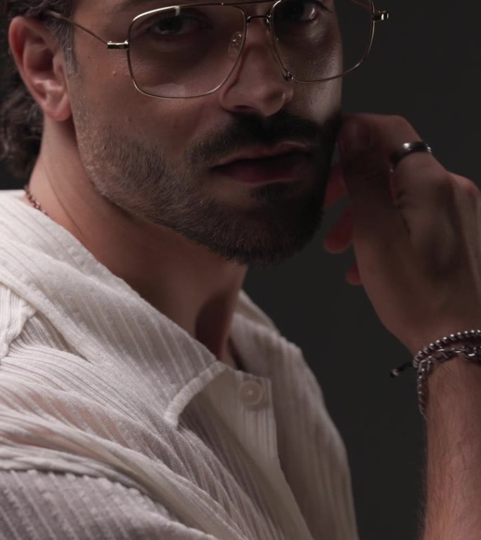侧视图视频，性感的大胡子男子带着眼镜看向一边，触摸脸，在灰色背景的闪光灯前很有诱惑力