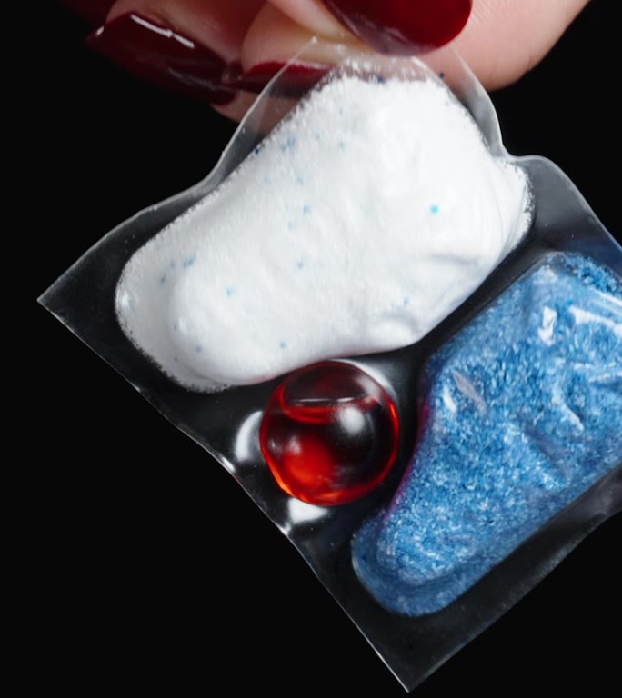 垂直视频。一个女人手里拿着一个可溶的小包，里面装着蓝白相间的洗碗粉和红色的凝胶。它被隔离在黑色的背景
