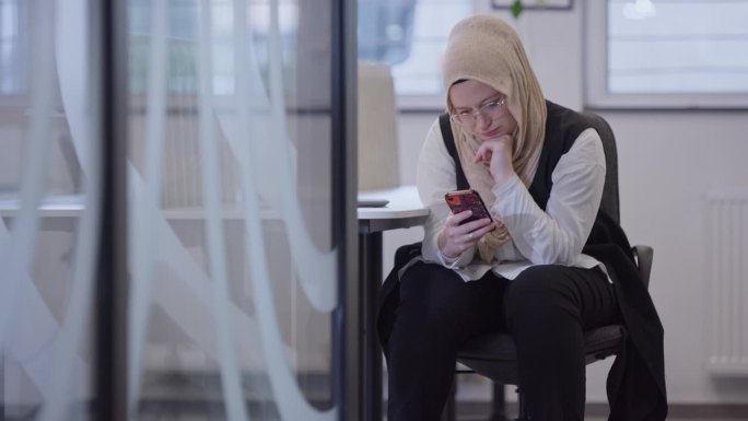 一位戴着头巾的女商人在现代化的办公室里休息，在现代的工作空间里使用她的智能手机，突出了企业界的多样性