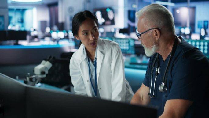 现代医院研究实验室中，亚洲女科学家使用台式电脑与白人男医生交谈。同事讨论病人的检查结果、治疗方法、药