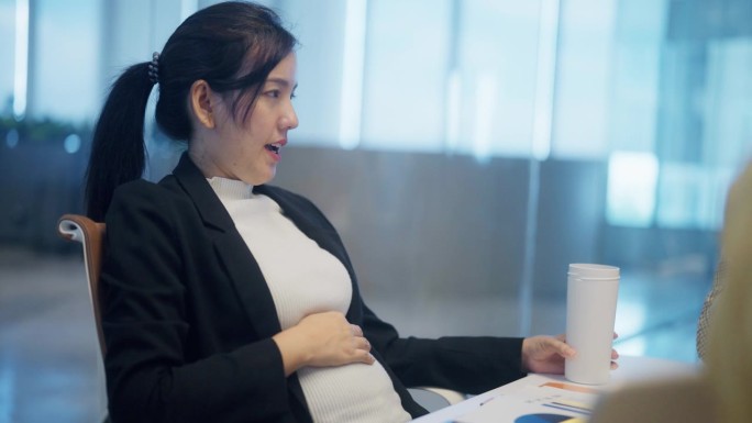 亚洲怀孕经理坐在桌边的侧视图在办公室工作时头脑风暴和揉肚子。