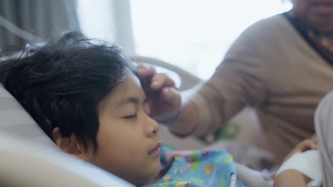 一位亚洲祖母抚摸着一个躺在医院病床上的小男孩的头，她表达了她的爱，并希望他早日康复。