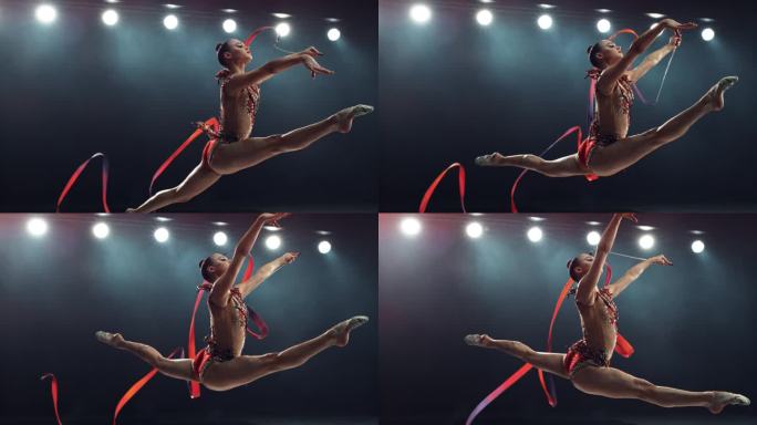 灯光大舞台上的现代艺术体操表演。美丽的女运动员在用红丝带创造形状的同时做出完美的分裂跳跃。超级慢动作