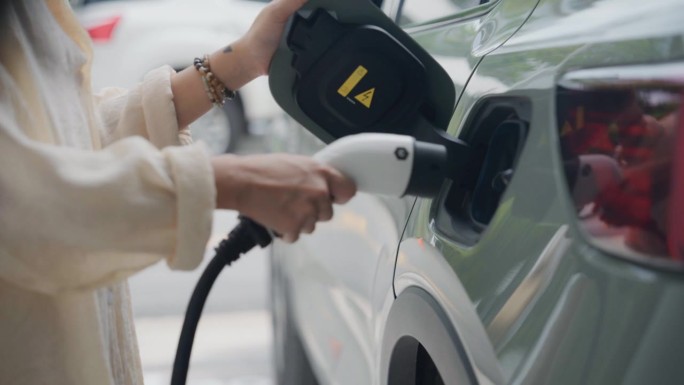 一名亚洲妇女将电动汽车插头插入汽车进风口，在有充电站的停车场充电。