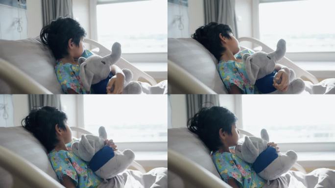 周末，一个年轻的亚洲病人抱着一个娃娃躺在医院的病床上，望着窗外。