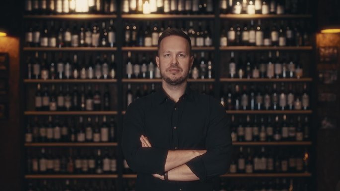 一位成年男性餐厅老板的肖像，背景是一家高档餐厅的酒架上。侍酒师可以提供精品葡萄酒从存储设施。