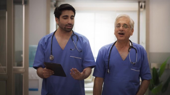 印度年轻和资深男医生和护士在医院走廊使用数字平板电脑和谈话的慢动作肖像。医学专家分享关于病人手术的笔
