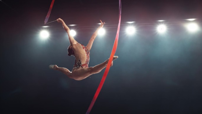 优雅的艺术体操运动员用丝带做一个有力的劈叉跳跃。年轻女性穿着漂亮的制服在黑暗的房间里锻炼。电影超级慢
