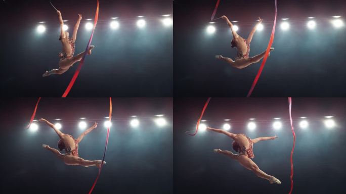 优雅的艺术体操运动员用丝带做一个有力的劈叉跳跃。年轻女性穿着漂亮的制服在黑暗的房间里锻炼。电影超级慢