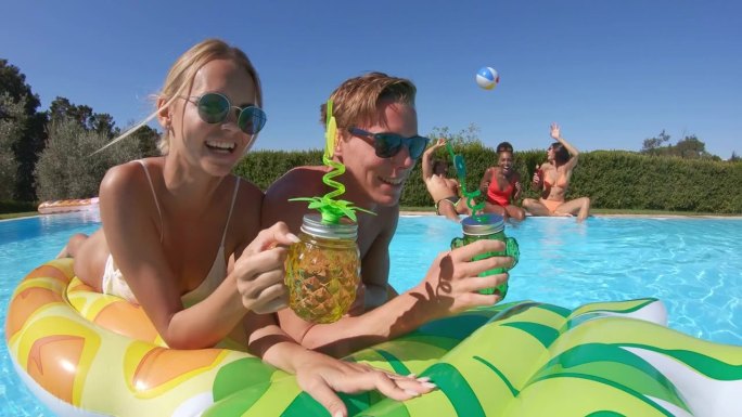 身着泳装的多民族青年朋友在泳池边欢度夏日的写真。一群年轻人在阳光明媚的日子里在户外喝鸡尾酒