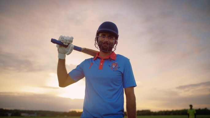 放大一名南亚板球运动员的肖像，穿着蓝色制服，戴着防护头盔站在球场上。专业的印度运动员看着镜头，微笑着
