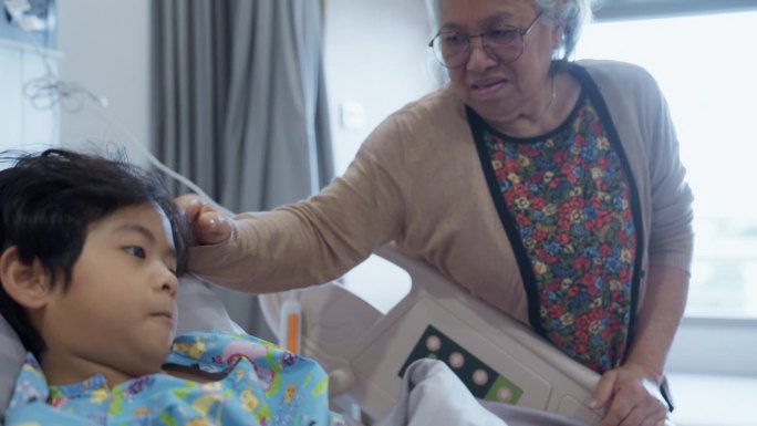一位亚洲祖母抚摸着躺在病床上的小孩的头，她在病床旁表达她的爱。