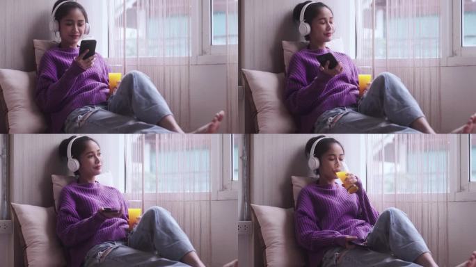 快乐的年轻亚洲女子坐在窗边，喝着橙汁，在家通过手机和耳机听音乐。