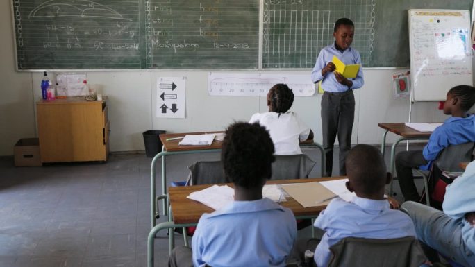 特写镜头。在非洲的一间教室里，一个非洲黑人学生站起来给全班同学读书