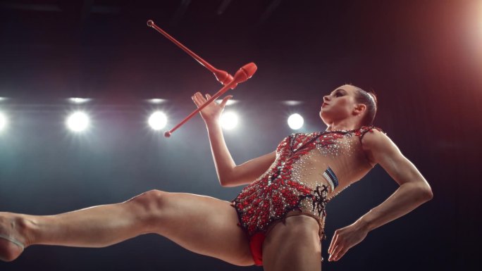 才华横溢的年轻艺术体操运动员用红色球杆表演一套常规动作。在黑暗的舞台上，穿着制服的年轻女性拿着棍棒杂