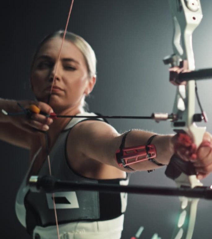 垂直屏幕:一名女弓箭手用现代复合弓瞄准目标射箭的电影肖像。强壮的运动员装备齐全，并配有胸部和手臂防护