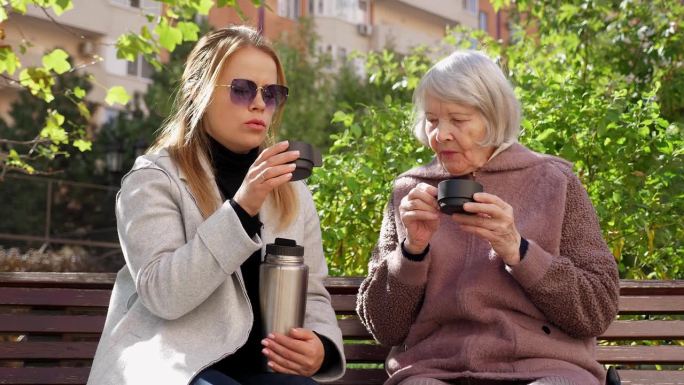 一位成年妇女和一位年老的祖母坐在公园的长椅上喝茶。