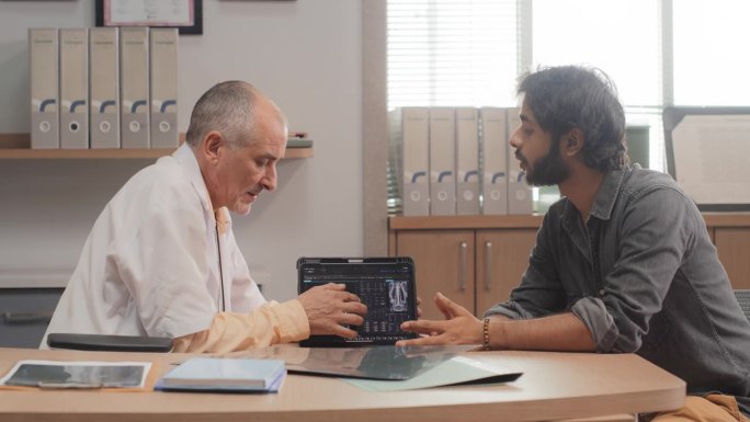 医院病房:印度中年男医生在预约期间与病人交谈，展示并解释电子平板电脑上的脑部CT MRI扫描结果。医