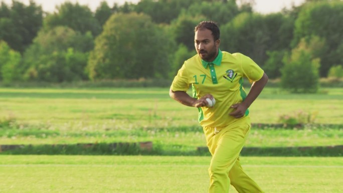 一名穿着黄绿制服的南亚板球运动员在球场上投球的慢动作肖像。职业印度投球手是专注的，目标是击中三柱门与