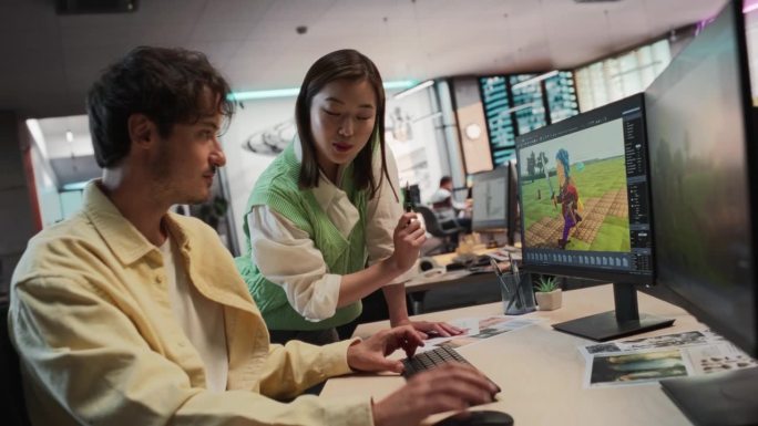 白人男性游戏设计师使用桌面电脑和3D建模软件，与游戏开发工作室办公室的亚洲女性艺术总监交谈。不同的同