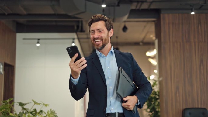 穿着正式服装的时髦男老板悠闲地走在联合办公空间里，用智能手机看搞笑视频。微笑的男人手里拿着黑色文件夹