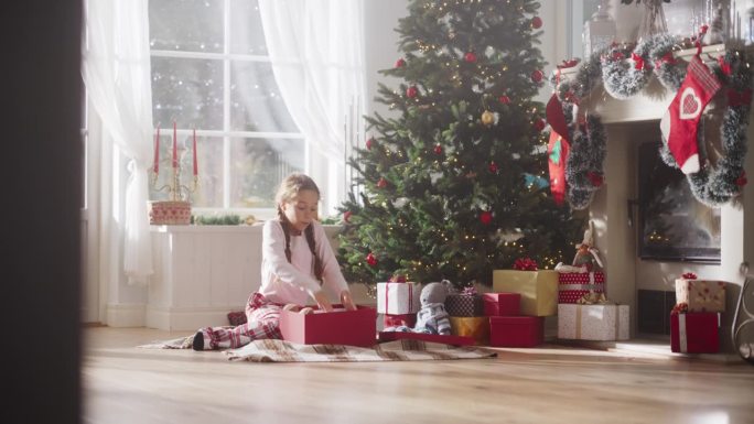 快乐的小女孩在节日早晨醒来，从圣诞树下收到新玩具的慢镜头。可爱的孩子迫不及待地从圣诞老人那里得到礼物