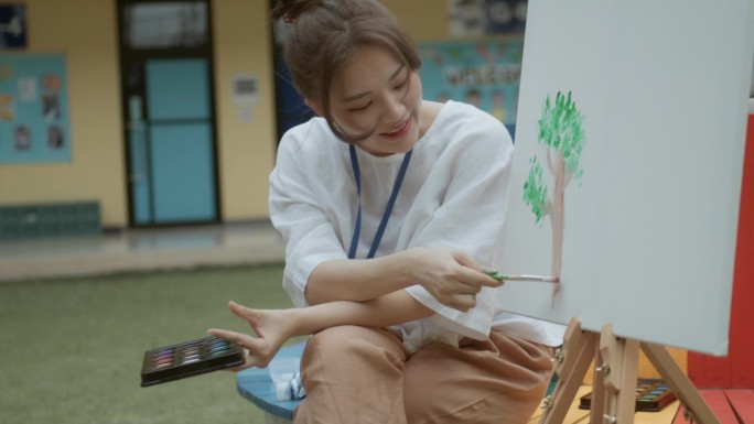 美丽的亚洲老师教幼儿园小朋友坐在教室外面用水彩画。