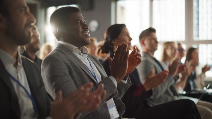 一名非洲男子坐在科技会议拥挤的礼堂里。黑人商人听主题演讲。专家观看创新技术展示并与观众鼓掌。慢动作