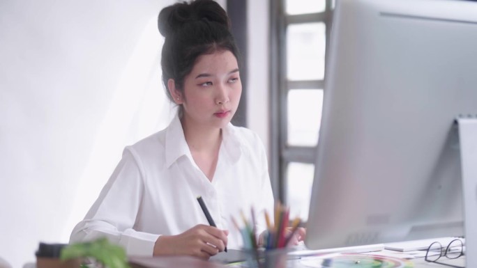 年轻的亚洲女性自由平面设计师或艺术家修图师，用触控笔在数字平板电脑上画画，看着电脑屏幕显示器，在她的