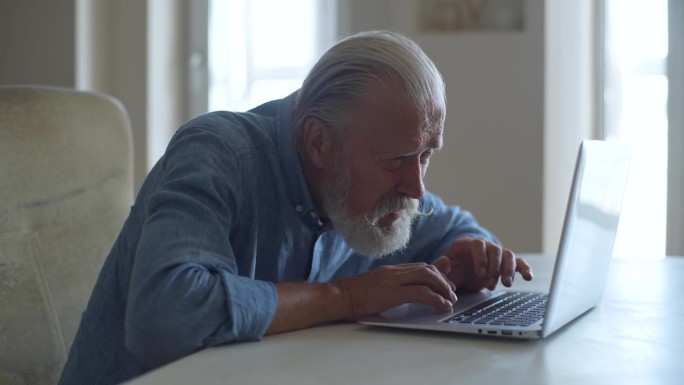 侧视图，年长的成年男性坐在笔记本电脑前，看着屏幕，脸上带着担忧的表情。专注的成熟男子看着电脑屏幕