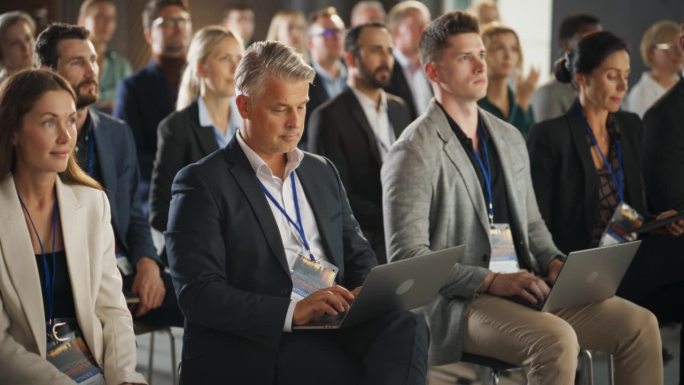 在医学峰会上，白人男性制药公司CEO坐在不同的人群中，使用笔记本电脑。成功人士聆听有关生物制药和医学