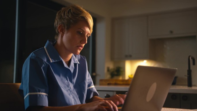 穿着护士服的女人晚上在家里用手机工作或用笔记本电脑学习
