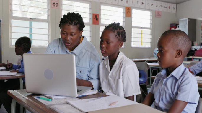 特写镜头。在非洲的一间教室里，非洲黑人女教师坐在桌前，用笔记本电脑帮助非洲孩子完成任务