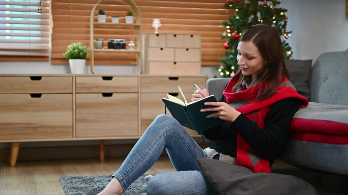 迷人的年轻女子坐在客厅里，在美丽的圣诞树前，在个人日常计划表上写笔记。