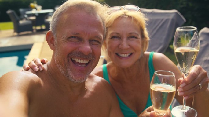 老年夫妇在度假时摆姿势自拍，喝着香槟，在游泳池边放松