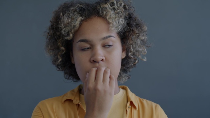 紧张的年轻非洲裔美国妇女咬指甲感到焦虑的特写慢动作肖像在灰色背景