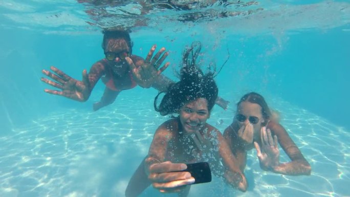女友用智能手机在水下自拍的慢镜头。跟踪拍摄三个女人在炎热的夏天享受茶水，在水疗中心或酒店游泳
