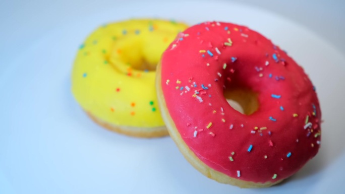 两个甜甜圈在白色的背景上旋转，上面有黄色和粉色的釉面，还有香蕉和草莓口味的彩色糖屑。