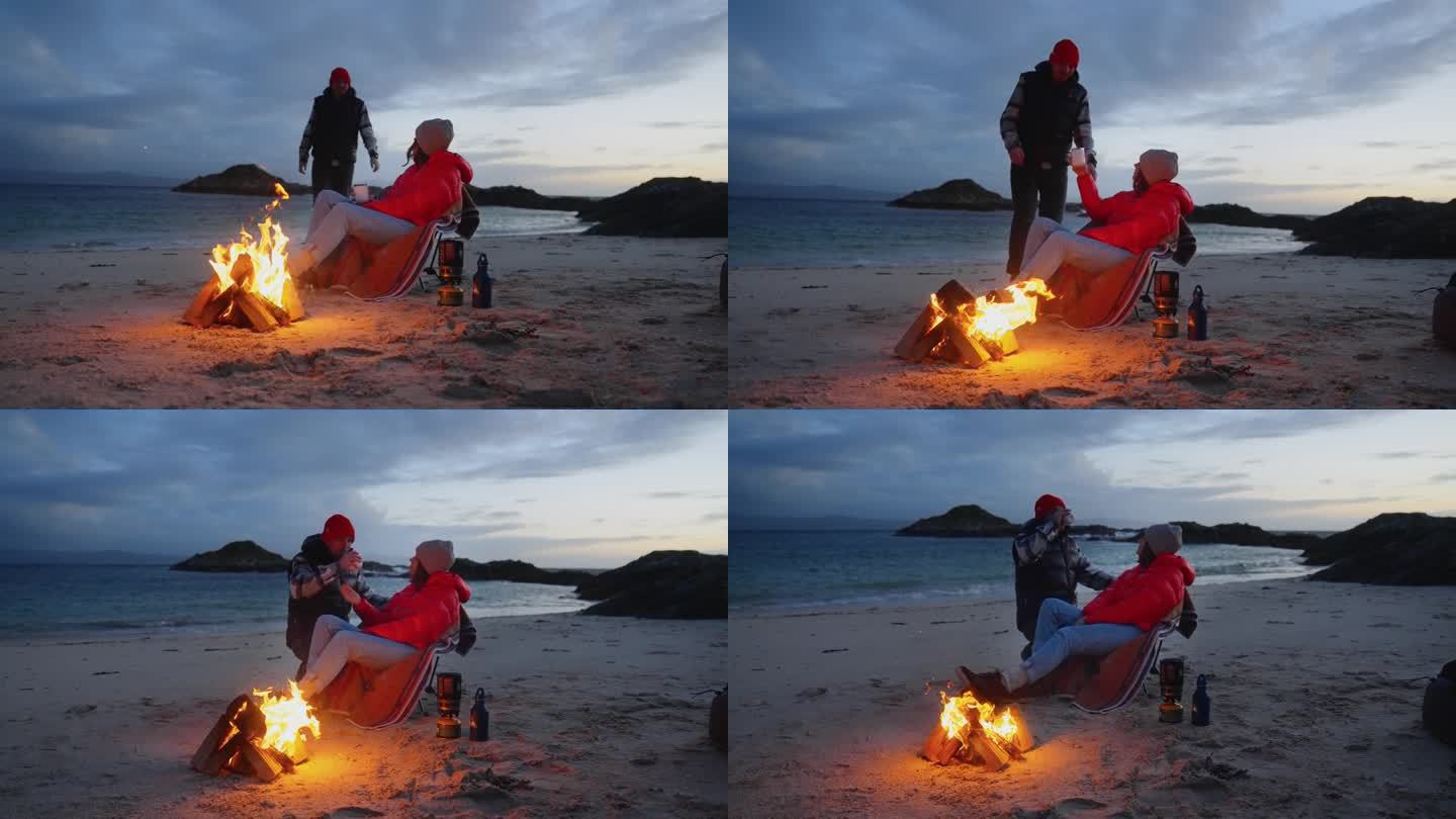 一对浪漫的夫妇穿着暖和的衣服在寒冷的天气里露营。一起在沙滩上篝火边热身