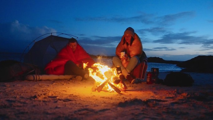 一对浪漫的夫妇穿着暖和的衣服在寒冷的天气里露营。一起在沙滩上用营火取暖，喝茶