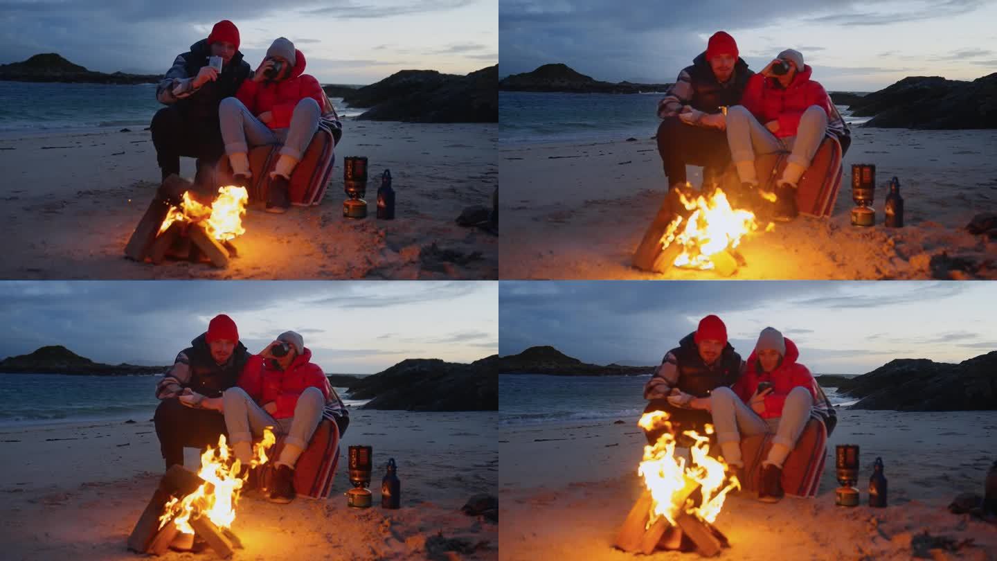 一对浪漫的夫妇穿着暖和的衣服在寒冷的天气里露营。在篝火旁喝热茶暖身