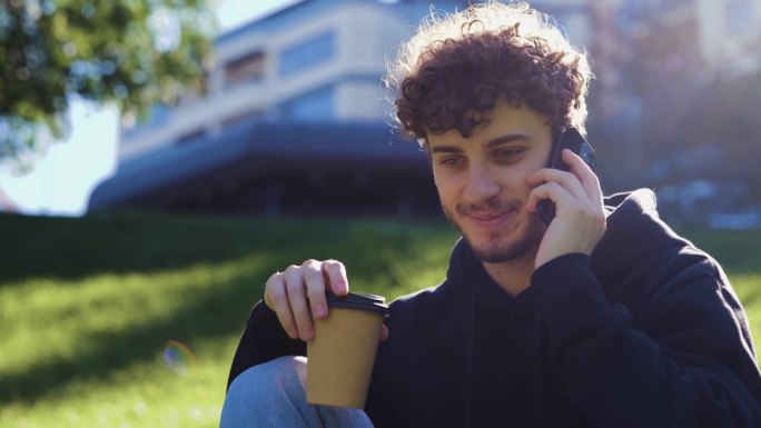在城市的公园里，一个快乐的年轻人一边喝咖啡一边用智能手机聊天。的生活方式。秋季散步的概念。实时概念。
