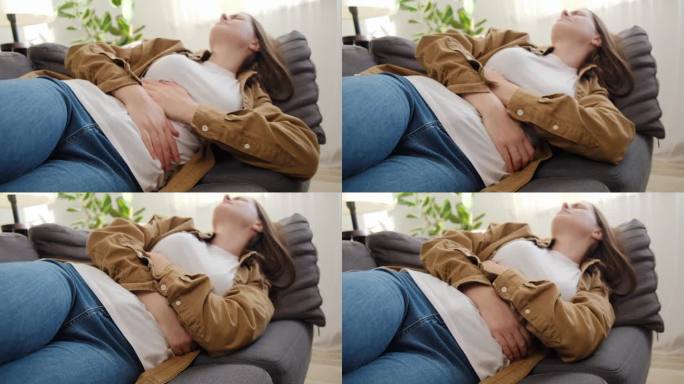 患有月经疼痛的患病年轻女性的选择性焦点。悲伤的女孩用手捏着肚子，胃疼或痛经，躺在家里的沙发上。腹痛概