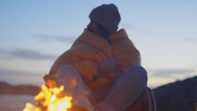 一对浪漫的夫妇穿着暖和的衣服在寒冷的天气里露营。裹着厚厚的毯子，在沙滩上围着篝火取暖