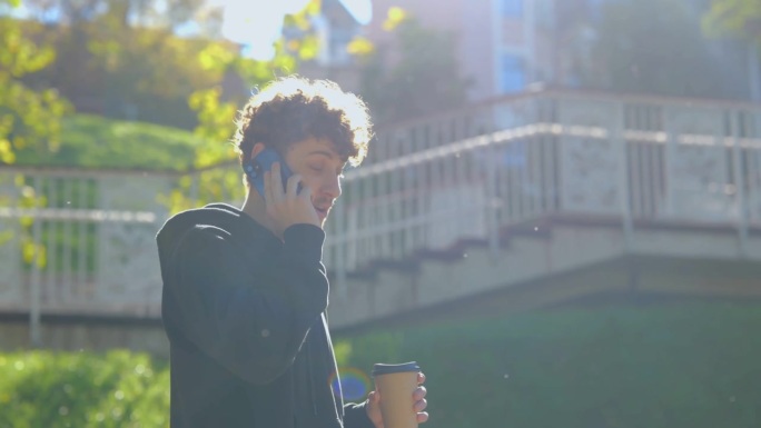 快乐的年轻人一边在公园散步一边拿着咖啡一边讲手机。的生活方式。秋季散步的概念。实时概念。