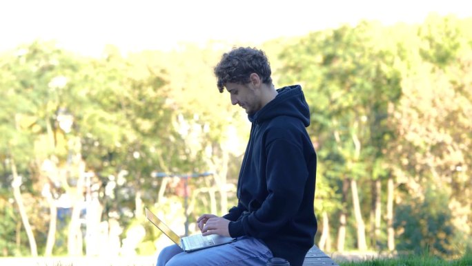 微笑的男人一边喝咖啡，一边坐在公园的长椅上，一边用笔记本电脑工作。秋天的季节。教育，工作理念。实时概